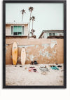 Een strandtafereel met twee surfplanken die tegen een betonnen muur leunen met een verweerd uiterlijk, dat lijkt op een Surfbord Op Het Strand Schilderij. Kleding en persoonlijke spullen worden op het zand voor de muur neergelegd. Op de achtergrond zijn twee huizen en hoge palmbomen zichtbaar, waardoor een perfecte wanddecoratie van CollageDepot ontstaat.,Zwart-Zonder,Lichtbruin-Zonder,showOne,Zonder