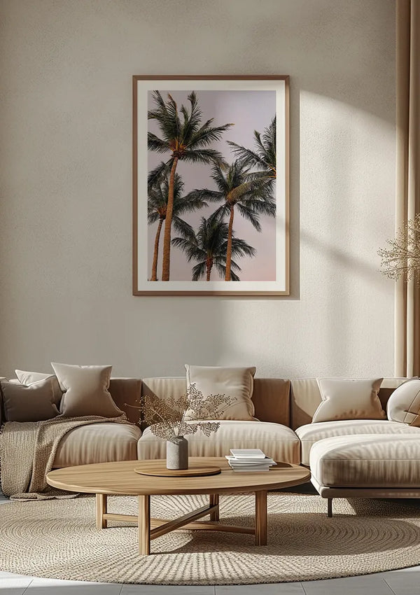 Een minimalistische woonkamer met beige muren met een groot ingelijst schilderij Hoge Palmbomen van CollageDepot dat boven een beige sectionele bank met kussens hangt. Voor de bank staat een ronde houten salontafel met een vaasje en gedroogde bloemen. De wanddecoratie draagt bij aan de serene sfeer van de ruimte.,Lichtbruin
