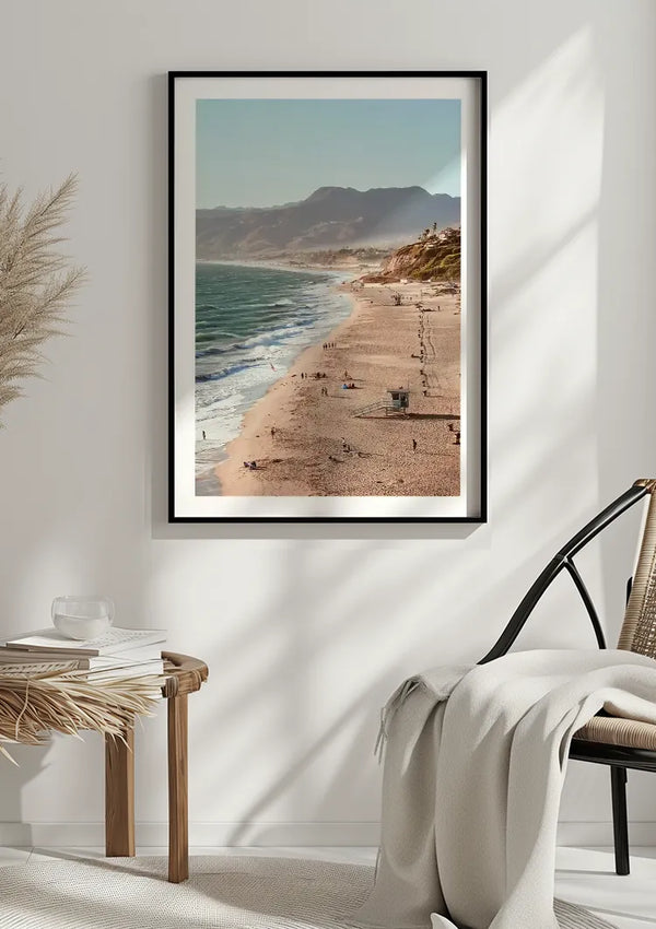 Een ingelijste foto van een strandtafereel aan de kust, of het Geweldige Uitzicht Schilderij van CollageDepot, hangt aan een witte muur. Het strand heeft golven, zand en bergen op de achtergrond. Onder de foto staat een rieten kruk met boeken en een glas. Een aangrenzende zwarte stoel is gedrapeerd met een witte deken.,Zwart