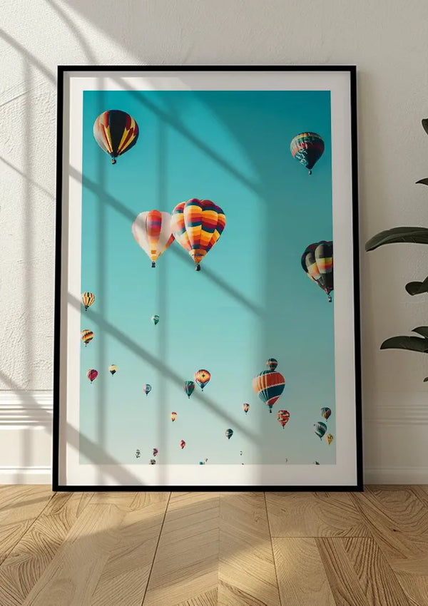 Deze boeiende CollageDepot wanddecoratie is een ingelijste foto die tegen een witte muur leunt en toont het kleurrijke Vele Luchtballonnen Schilderij zwevend in een helderblauwe lucht. Het zonlicht werpt schaduwen op de muur en de houten vloer. Rechts is gedeeltelijk een plant met groene bladeren zichtbaar.,Zwart