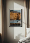 Een ingelijst Surfen bij kliff schilderij van CollageDepot hangt aan een beige muur naast witte gordijnen. De foto toont een strandtafereel met een persoon die in ondiep water staat, met uitzicht op een grote klif op de achtergrond. Het zonlicht stroomt de kamer binnen en werpt schaduwen op de muur en het bed.