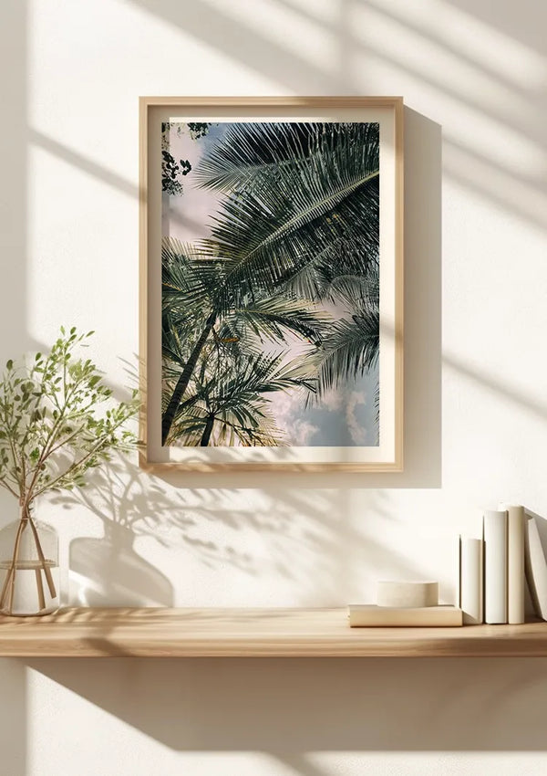 Aan een witte muur hangt een ingelijste foto van palmbladeren tegen een hemel, beveiligd door een magnetisch ophangsysteem. Op een houten plank onder het Palmboom Bladeren Schilderij van CollageDepot staat een klein groen plantje in een vaas en een paar gestapelde boeken. Natuurlijk licht werpt schaduwen op de muur en plank.,Lichtbruin