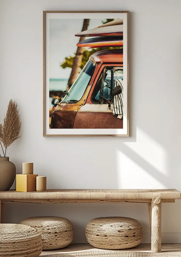 Aan een lichtgekleurde muur boven een minimalistische houten bank hangt een ingelijste foto van een vintage voertuig met een surfplank erop. De bank heeft eronder twee geweven kussens en enkele decoratieve items, waaronder een vaas met gedroogde planten en gestapelde kaarsen. Deze wanddecoratie, het CollageDepot Busje Met Surfborden Schilderij, is voorzien van een handig magnetisch ophangsysteem.,Lichtbruin