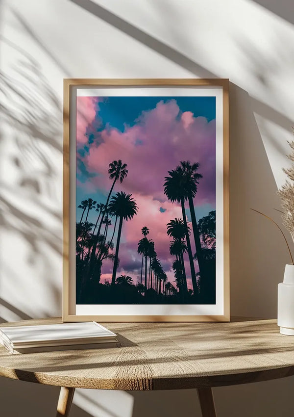 Een ingelijste foto van hoge palmbomen, afgetekend tegen een levendige roze en blauwe zonsondergang, staat op een houten tafel en dient als prachtige wanddecoratie. Naast het frame zijn verschillende gesloten boeken gestapeld en op de muur erachter zijn schaduwen van raamkozijnen en planten geworpen. Het product heet Roze Wolken Schilderij van CollageDepot.,Lichtbruin