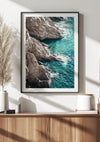 Aan een witte muur hangt een ingelijste foto van een rotsachtige kustlijn met turquoise water en dient als perfecte wanddecoratie. Het Rotsachtig Schilderij van CollageDepot zet een betoverende toon voor de kamer. Beneden staat in een houten kast een witte vaas met gedroogd pampagras, een kaars, een klein boekje en een paar potten. Het zonlicht stroomt naar binnen en werpt schaduwen.,Zwart