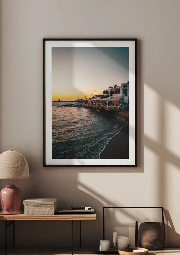 Een groot ingelijst CollageDepot Restaurantjes Aan De Kust Schilderij van een zonsondergang aan het water is op een beige muur gemonteerd. Onder het frame staat een tafeltje met een beige tafellamp, een stapel boeken en decoratieve voorwerpen. Zonlicht werpt schaduwen op de muur en vloer, waardoor een warme sfeer ontstaat.,Zwart