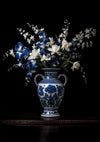 Een gedetailleerd bloemstuk in een blauw-witte porseleinen vaas wordt weergegeven tegen een zwarte achtergrond. Het boeket bestaat uit een mix van witte en blauwe bloemen met groene bladeren, symmetrisch gerangschikt. Met ingewikkelde blauwe patronen en twee handvatten lijkt de vaas op een elegant wit en blauwe bloemen in het donker schilderij van CollageDepot.