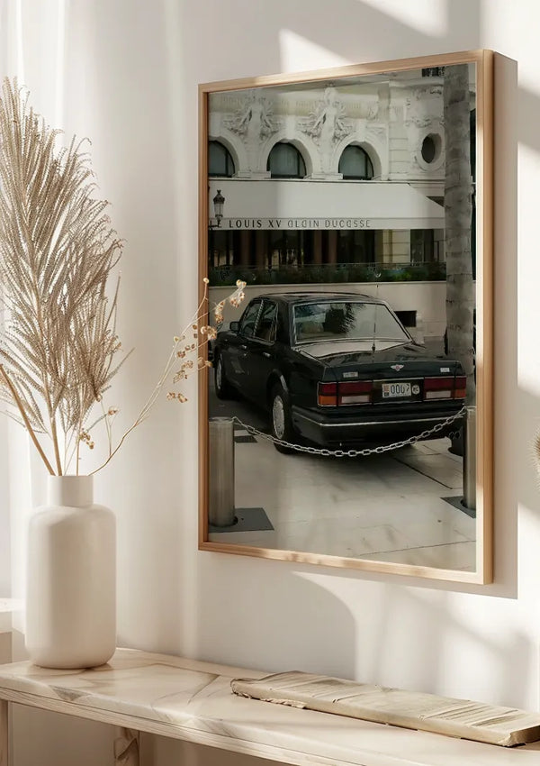 Een ingelijste spiegel of glazen paneel weerspiegelt een buitenscène met een klassieke zwarte auto geparkeerd nabij een kettingversperring. Op de achtergrond staat een wit gebouw met een bord met de tekst "Lodewijk XV Alain Ducasse." Binnen is links een vaas met gedroogde planten zichtbaar. Dit elegante Klassieke Bentley Auto Schilderij van CollageDepot voegt charme toe aan elke ruimte.,Lichtbruin