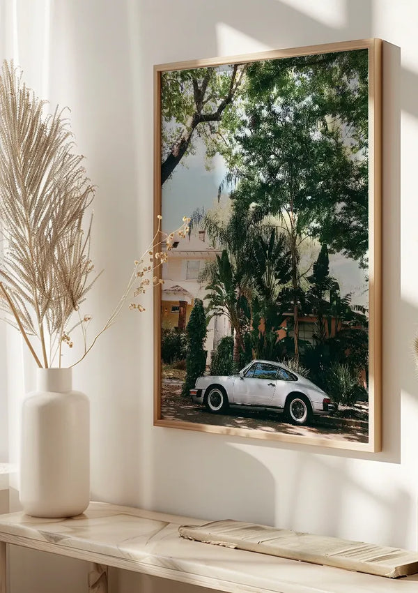 Een ingelijste foto aan de muur, waarop een vintage witte auto is vastgelegd die doet denken aan een Porsche 911, is het perfecte Zilveren Porsche 911 Oldtimer Schilderij van CollageDepot. Het serene tafereel toont de auto die geparkeerd staat voor een huis, omgeven door weelderig groen en bomen. Hangend boven een lichtgekleurde houten consoletafel met twee siervazen en gedroogde planten, vastgezet door een magnetisch ophangsysteem.,Lichtbruin