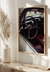 Aan een beige muur hangt een ingelijste foto van het interieur van een klassieke Ferrari en dient als prachtige wanddecoratie. De focus ligt op het stuur met het iconische Ferrari-logo. Zacht zonlicht verlicht het beeld en de omringende beige vaas en pampasgras, alles boven een netjes gerangschikte plank. Dit prachtige stuk staat bekend als Steering Wheel Ferrari Schilderij van CollageDepot.,Lichtbruin