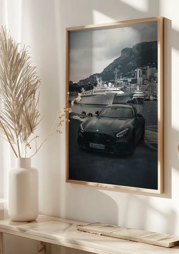 Aan een witte muur hangt een ingelijste foto van een zwarte auto, waarop een luxe auto is afgebeeld, geparkeerd aan de waterkant met jachten en de bergachtige achtergrond van Monaco. Zonlicht verlicht de scène en werpt schaduwen op nabijgelegen gedroogde planten en een lichtgekleurde plank, waardoor een prachtige wanddecoratie ontstaat: het Mercedes AMG GTR Monaco Schilderij van CollageDepot.,Lichtbruin