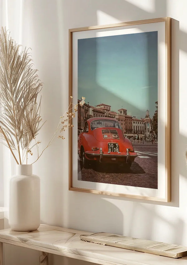 Een ingelijste foto aan de muur toont een rode klassieke auto met een Italiaans kenteken, geparkeerd op straat. De achtergrond toont historische gebouwen onder een heldere hemel. Een witte vaas met gedroogde planten staat op een lichtgekleurde houten plank onder het **Klassieke Rode Porsche 356 Schilderij** van **CollageDepot**, beveiligd door een magnetisch ophangsysteem.,Lichtbruin