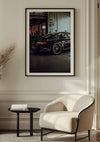 Een ingelijste foto van een zwarte Porsche GT3 In Een Garage van CollageDepot is met behulp van een magnetisch ophangsysteem aan een beige muur gemonteerd. Beneden staat een moderne beige fauteuil naast een klein rond zwart bijzettafeltje met een stapel boeken. De setting is een stijlvolle, minimalistische kamer.,Zwart