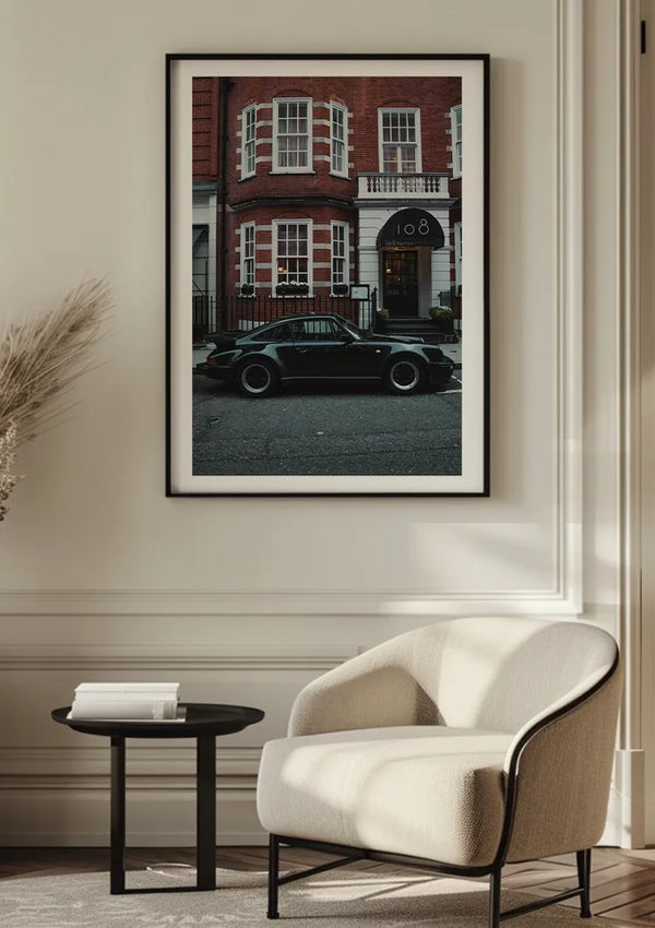 Een ingelijst CollageDepot Zwart Klassieke Porsche Turbo Schilderij aan de muur toont een zwarte Porsche Turbo, geparkeerd voor een bakstenen gebouw met een witte deur en het nummer "108" boven de ingang. De kamer is voorzien van een witte moderne stoel en een donkere ronde bijzettafel, geplaatst tegen gedecoreerde kunst aan de muur opgehangen met een magnetisch ophangsysteem.,Zwart