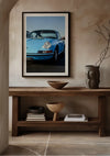 Een ingelijst CollageDepot Vintage Porsche 911 schilderij hangt boven een houten consoletafel. Op de tafel staan decoratieve voorwerpen, waaronder een grijze vaas met takken, een keramische kom en gestapelde boeken. De neutrale, aardse tinten van de setting vullen de verfijnde wanddecoratie perfect aan.,Zwart