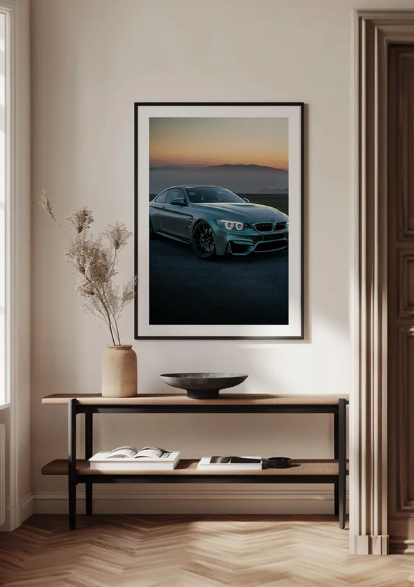 Een ingelijste foto van een BMW M3 Bij zonsondergang Schilderij van CollageDepot hangt aan een witte muur boven een houten consoletafel. Op de tafel staat een decoratieve vaas met gedroogde planten, een open boek en een ondiepe schaal. De setting oogt minimalistisch en overzichtelijk, versterkt door het magnetische ophangsysteem voor naadloze wanddecoratie.,Zwart