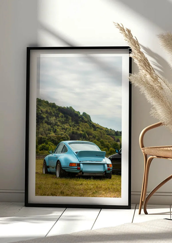Een ingelijste foto van een Babyblauw Porsche 911 Schilderij, geparkeerd op een grasveld, staat tegen een lichtgekleurde muur. De foto, die gebruik maakt van een magnetisch ophangsysteem, leunt naast een rieten stoel en pampasgras in de lichte, zonovergoten kamer – een perfecte keuze voor wanddecoratie van CollageDepot.,Zwart