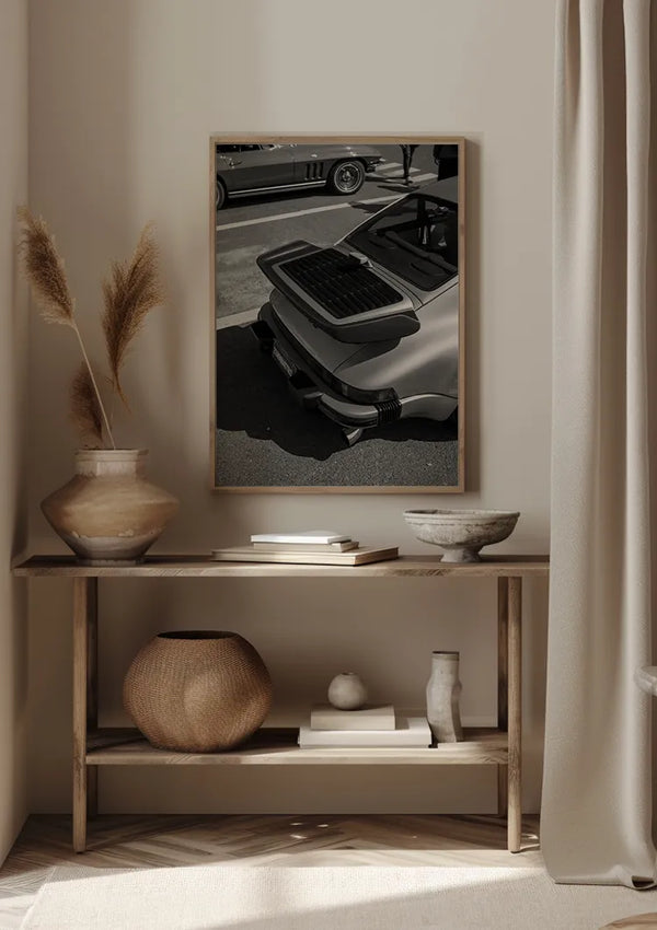 Een minimalistische kamer met een houten consoletafel tegen een beige muur. Op de tafel staan decoratieve voorwerpen, waaronder vazen en een mand. Boven de tafel hangt een ingelijste zwart-witfoto van de achterkant van een klassieke auto, het Rear View Porsche Carrera Schilderij van CollageDepot, aan de muur, beveiligd met een elegant magnetisch ophangsysteem voor eenvoudige plaatsing.,Lichtbruin