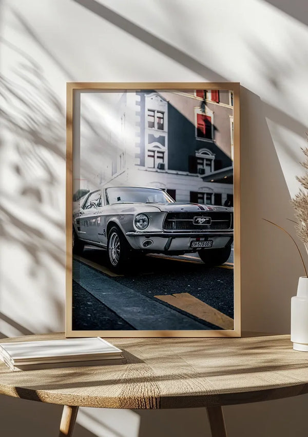 Ingelijste foto van een vintage zilveren Ford Mustang, geplaatst op een houten oppervlak. Zonlicht en schaduwen van een raam voegen textuur toe aan de scène. De achtergrond bestaat uit een stadsstraat met gebouwen, wat de klassieke uitstraling van de auto versterkt: echt een CollageDepot Klassieke Mustang Schilderij voor elke ruimte.,Lichtbruin