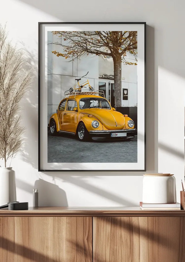 Ingelijste wanddecoratie toont een felgele vintage Volkswagen Kever met een fiets op het dak, geparkeerd naast een boom en een modern gebouw. Dit Gele Volkswagen Kever Schilderij van CollageDepot, opgehangen boven een houten kast met decoritems, voegt charme toe aan elke kamer. De ingelijste foto is voorzien van een eenvoudig magnetisch ophangsysteem.,Zwart