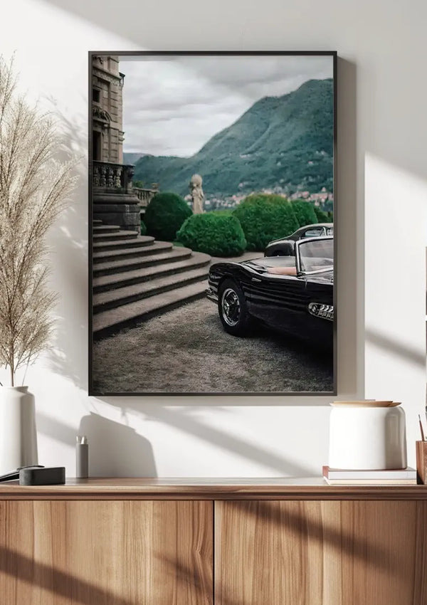 Een ingelijste foto aan de muur toont een vintage zwarte BMW-cabrio geparkeerd naast de stenen trap die naar een klassiek gebouw leidt. Op de achtergrond zijn groene bergen onder een bewolkte hemel zichtbaar. Dit elegante Klassieke BMW Cabrio Schilderij van CollageDepot hangt boven een houten kast met decoratieve items.,Zwart