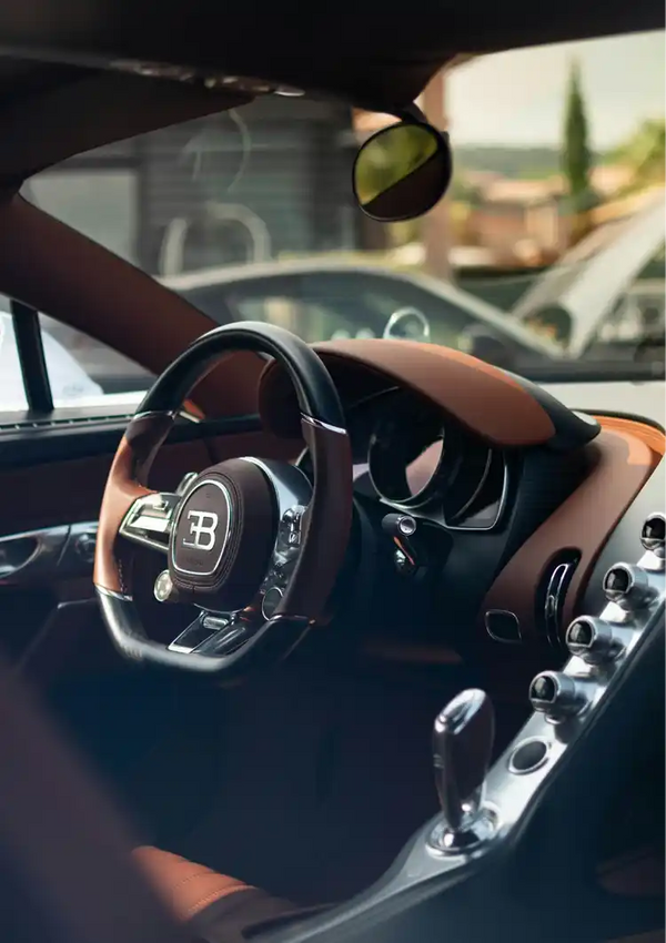 Het interieur van een luxe auto met een gedetailleerd stuur met het Bugatti-logo, een verfijnd dashboard met verschillende bedieningselementen en een strakke versnellingspook. Het uitzicht omvat ook de achteruitkijkspiegel en een deel van het raam, allemaal ingelijst als een Bugatti Chiron Interior Schilderij van CollageDepot, dat luxe wanddecoratie biedt voor autoliefhebbers.