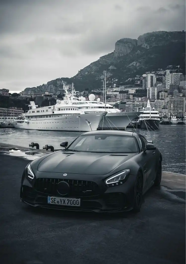 Een matzwarte sportwagen, of zwarte auto, staat geparkeerd bij een jachthaven met op de achtergrond enkele luxe jachten. Het tafereel, dat doet denken aan Monaco, speelt zich af tegen een bewolkte hemel met een stadsgezicht en een heuvel in de verte. Deze schilderachtige omgeving zou een prachtig Mercedes AMG GTR Monaco Schilderij van CollageDepot wanddecoratie opleveren.-