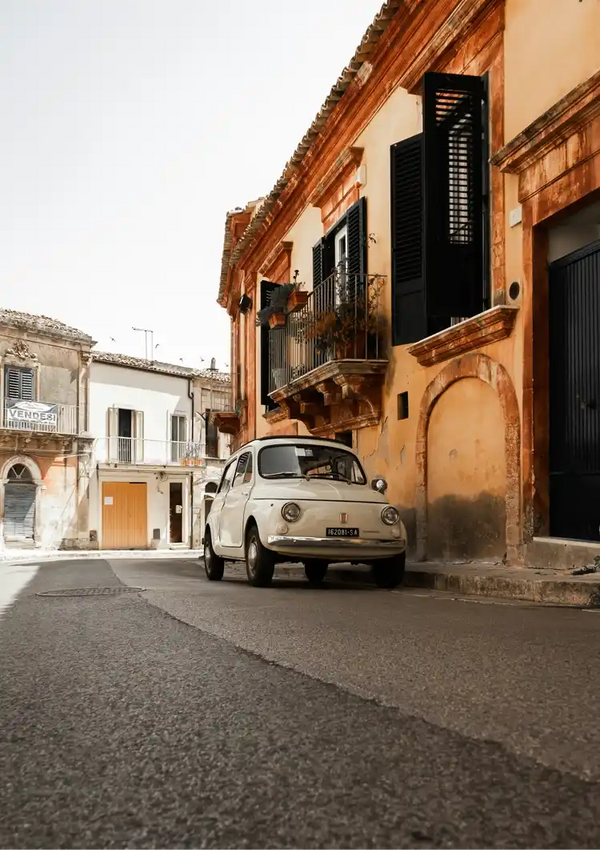 Een Witte Vintage Fiat 500 Schilderij van CollageDepot staat geparkeerd in een smal straatje vol oude gebouwen met houten luiken en balkons. De scène wordt verlicht door daglicht en de omgeving lijkt rustig zonder zichtbare mensen, waardoor het aanvoelt als een pittoreske wanddecoratie.-