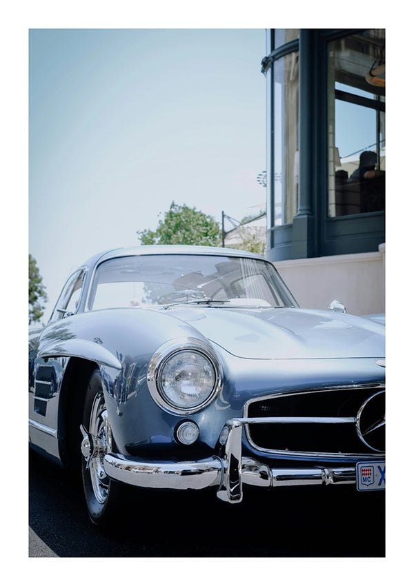 Een laaghoekaanzicht van een vintage zilveren Mercedes-Benz 300SL geparkeerd op straat. De auto heeft een glanzende chromen grille en ronde koplampen, waardoor het bijna lijkt alsof hij deel uitmaakt van een elegant CollageDepot Mercedes 300SL Schilderij. Er is een gebouw met grote ramen en bomen op de achtergrond.-