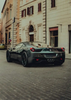 Een zwarte Ferrari 488 In Oude Stad Schilderij van CollageDepot staat geparkeerd in een smalle geplaveide straat vol oude gebouwen. De auto heeft een strak, aerodynamisch design en opvallende achterlichten. De gebouwen hebben gebogen deuropeningen en ramen met luiken, die lijken op een scène uit een klassiek schilderij.-