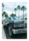 Een glanzend zwarte Chevy Caprice Schilderij van CollageDepot staat geparkeerd op straat naast een wit gebouw, met verschillende palmbomen en een blauwe lucht op de achtergrond. De auto beschikt over een chromen grille en grote chromen wielen, waardoor het net zo'n kunstwerk is als welke wanddecoratie dan ook.