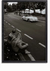 Een zwart-witfoto van een klassieke auto die over een verharde weg rijdt met bomen en toeschouwers op de achtergrond. Op de voorgrond zitten twee mensen met lang haar in winterkleding op de stoeprand en kijken naar de passerende auto. Dit nostalgische moment zou een perfecte Racende Porsche 356 Schilderij van CollageDepot voor je huis zijn.