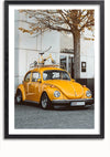 Een ingelijste foto toont een gele vintage Gele Volkswagen Kever Schilderij geparkeerd voor een modern gebouw. Op de imperiaal van de auto is een gele fiets gemonteerd. Er staat een bladloze boom vlakbij, wat een opvallend contrast geeft, perfect voor wanddecoratie met een magnetisch ophangsysteem van CollageDepot.,Zwart-Met,Lichtbruin-Met,showOne,Met