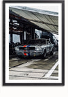 Een vintage zilveren raceauto versierd met rode en blauwe racestrepen staat geparkeerd onder een baldakijn, met vrachtwagentrailers en uitrusting op de achtergrond. De scène, die doet denken aan een iconisch Aston Martin-schilderij van CollageDepot, toont de auto klaar en klaar voor een evenement op het racecircuit.,Zwart-Met,Lichtbruin-Met,showOne,Met