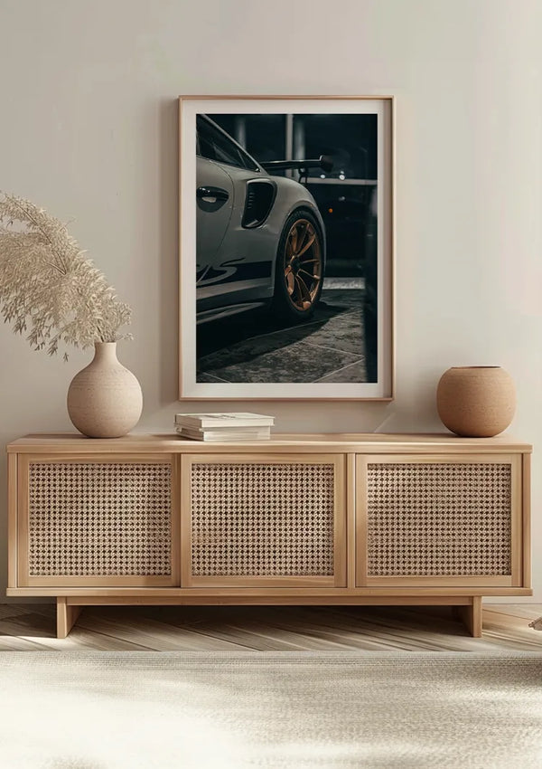 Een minimalistische woonkamer is voorzien van een lichthouten dressoir met drie rotandeuren. Bovenop staan twee ronde sierobjecten, een vaas met pampasgras en een stapel boeken. Een ingelijste foto van een Porsche GT3 RS-stuur is aan de muur boven het dressoir gemonteerd met behulp van een CollageDepot Striking Porsche GT3 RS Schilderij.,Lichtbruin