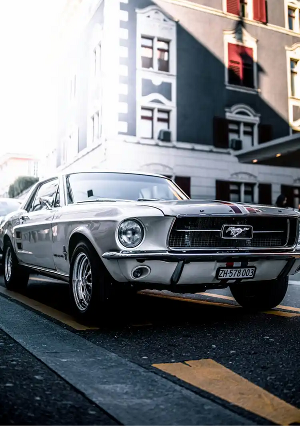 Een klassieke CollageDepot aaa 094 - auto geparkeerd in een stadsstraat, met een zilveren carrosserie en iconisch Mustang-embleem, met vage gebouwen op de achtergrond.-