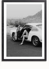 Een zwart-witfoto van een persoon die in een klassieke auto zit met de bestuurdersdeur open. De persoon draagt hoge hakken en de benen zijn zichtbaar buiten de auto. Dit opvallende beeld kan dienen als elegante wanddecoratie, waardoor het een tijdloos kunstwerk is voor elke kamer. De achtergrond toont een landschap met heuvels en schaarse vegetatie. Maak kennis met het Porsche With Love Schilderij van CollageDepot.,Zwart-Met,Lichtbruin-Met,showOne,Met
