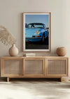 Een ingelijste foto van een vintage blauwe Porsche 911 is aan een beige muur gemonteerd boven een houten dressoir met rotan deuren. Het Vintage Porsche 911 Schilderij van CollageDepot wordt aangevuld met een beige vaas met gedroogd blad en een ronde terracotta vaas. Tussen de vazen rust een boek, wat de stijlvolle compositie versterkt.,Lichtbruin