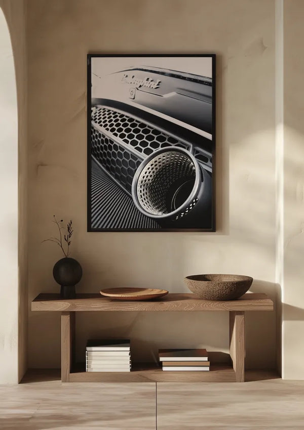 Een ingelijste foto met een close-up van een Lamborghini Huracan Evo-grille hangt aan een lichtgekleurde muur boven een houten consoletafel. Op de tafel staat een vaasje met takjes, een ondiepe schaal, een kom en verschillende gestapelde boeken. Zacht natuurlijk licht werpt subtiele schaduwen en creëert een elegante wanddecoratie met het Dream Exhaust Schilderij van CollageDepot.,Zwart