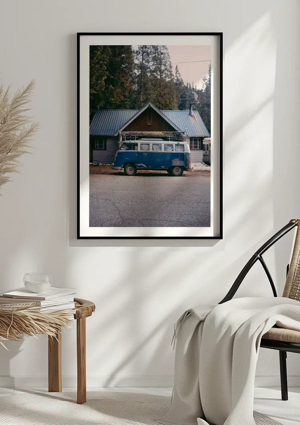 Op een witte muur wordt een ingelijste foto getoond van een Blauwe Volkswagenbus Schilderij van CollageDepot, geparkeerd voor een hut. De kamer bevat een rieten kruk met een stapel boeken en een kleine beker, een stoel met kussens en een gebreide plaid erover gedrapeerd. Deze charmante wanddecoratie geeft karakter aan de ruimte.,Zwart