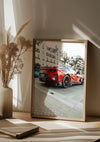 Een ingelijste foto van een rode Ferrari 812 Competizione, geparkeerd in een stadsstraat, wordt op een houten oppervlak geplaatst. De scène speelt zich af tegen het zonlicht dat door vitrages naar binnen valt. Links van het frame staat een beige keramische vaas met gedroogd gras, waardoor een elegant Ferrari 812 Competizione Schilderij van CollageDepot met magnetisch ophangsysteem ontstaat.,Lichtbruin