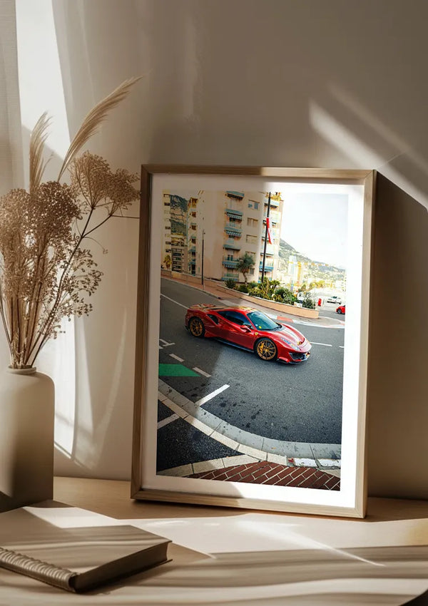 Een ingelijst CollageDepot Ferrari 488 Pista Monaco Schilderij met gouden randen wordt op een houten oppervlak geplaatst. Op de foto staat de auto op een bochtige weg nabij gebouwen. Naast het frame staan ook een vaas met gedroogde bloemstukken en een gesloten boek op het oppervlak, waardoor een elegante wanddecoratie ontstaat.,Lichtbruin