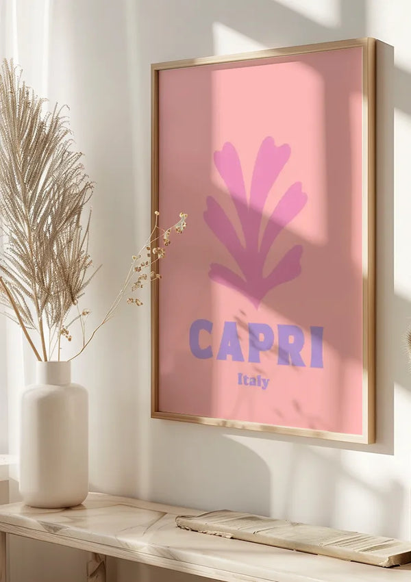 Een ingelijst Capri Italy-schilderij van CollageDepot hangt aan een muur en toont het woord "CAPRI" in opvallende paarse letters met "Italy" eronder. De roze achtergrond heeft een minimalistisch paars bloemenontwerp boven de tekst. Links maakt een vaas met gedroogde planten het tafereel compleet, waardoor dit minimalistische grafische schilderij wordt geperfectioneerd.,Lichtbruin