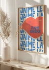 Een ingelijst J'aime Le Bonjour-schilderij van CollageDepot hangt aan een witte muur, met rode lippen waarop "J'aime Bonjour" staat geschreven. Op de achtergrond is de herhaalde blauwe tekst 'J'aime' te zien. Op een witte vensterbank naast het kunstwerk staat een witte vaas met gedroogde planten, die gebruik maakt van een magnetisch ophangsysteem voor eenvoudige presentatie.,Lichtbruin