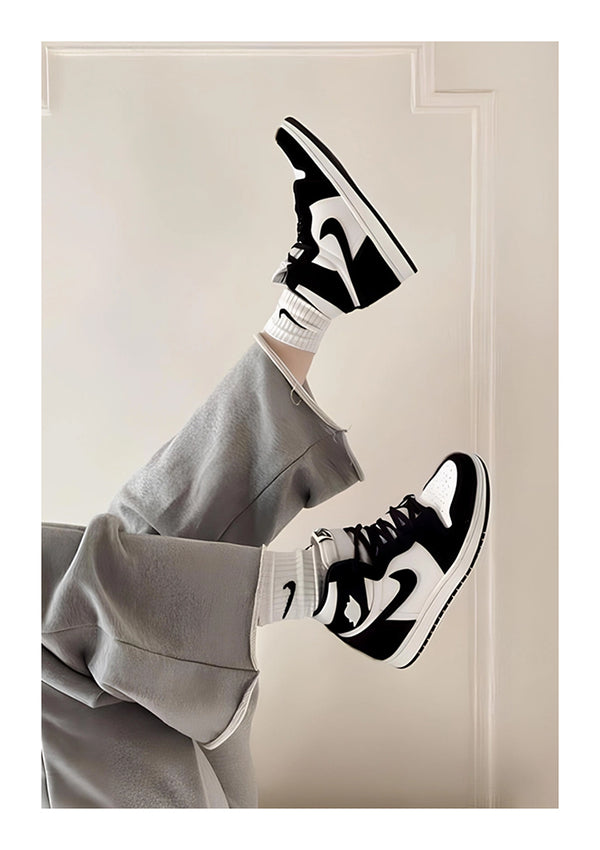 Er wordt een persoon gezien die achterover ligt met de benen omhoog in de lucht, gekleed in een grijze broek, witte sokken en zwart-witte Nike Jordans. De achtergrond is een neutraal gekleurde muur met paneeldetails en subtiele wanddecoratie met het Nike Jordans Schilderij van CollageDepot.-