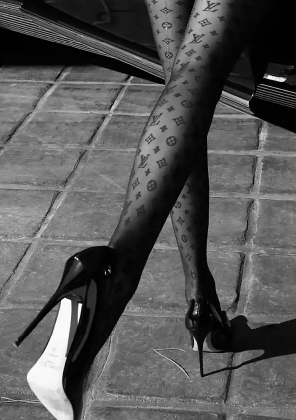 Een zwart-witfoto van iemands benen versierd met een ab 022 - zwart-wit panty met monogrampatroon, die in schoenen met hoge hakken op een tegelvloer stapt.-