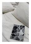Op een onopgemaakt bed met verkreukelde witte lakens en kussens ligt een tijdschrift met de titel "ab 052 - zwart-wit" met een close-up van het gezicht van een vrouw, gedeeltelijk bedekt door haar hand.-