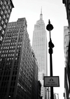 Een zwart-witfoto van een stadsgezicht met hoge gebouwen. Het hoogste gebouw, centraal gelegen, loopt taps toe naar een torenspits aan de bovenkant. Op de voorgrond is een straatlantaarn zichtbaar met een bord "busbanen foto afgedwongen". Perfect als wanddecoratie, dit Streets of New York schilderij van CollageDepot brengt stedelijke charme in elke ruimte.