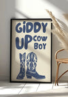 Op een ingelijste poster die tegen een muur leunt, staat de zinsnede "Giddy Up Cowboy" in vetgedrukte, gestileerde blauwe letters. Onder de tekst staat een illustratie van cowboylaarzen. Een rieten stoel en decoratief gedroogd pampagras zijn gedeeltelijk zichtbaar naast het Giddy Up Cowboy Schilderij van CollageDepot.,Zwart
