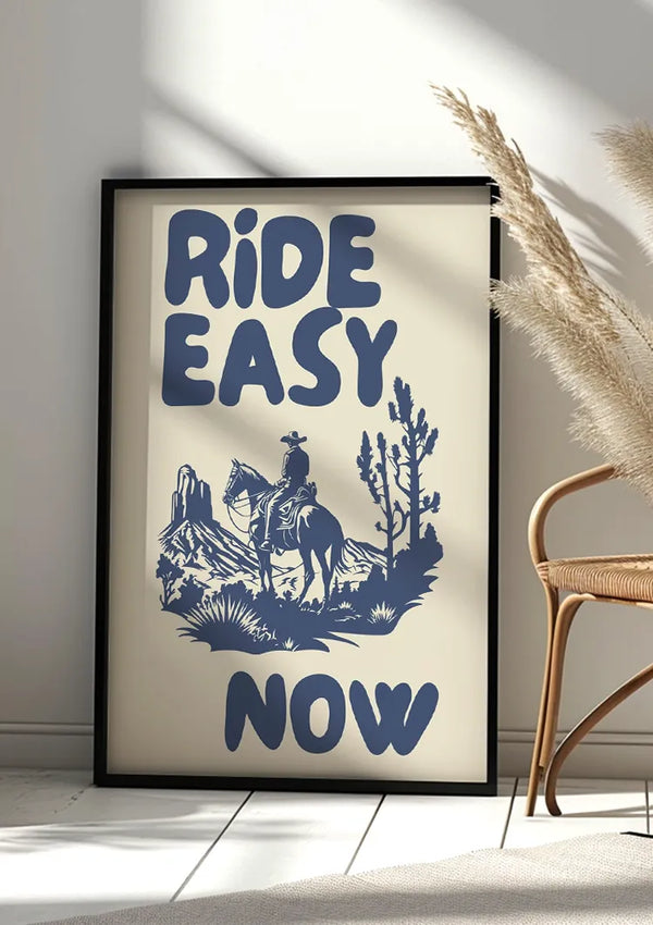 Tegen de muur staat een ingelijste poster met een blauwe illustratie van een cowboy op een paard tegen een woestijnachtergrond met de woorden "RIDE EASY NOW" in vetgedrukte letters. Dit is het Ride Easy Now-schilderij van CollageDepot. Aan de rechterkant zijn een rieten stoel en pampasgras te zien. Het tafereel, dat doet denken aan een westers landschap, wordt verlicht door natuurlijk zonlicht.,Zwart